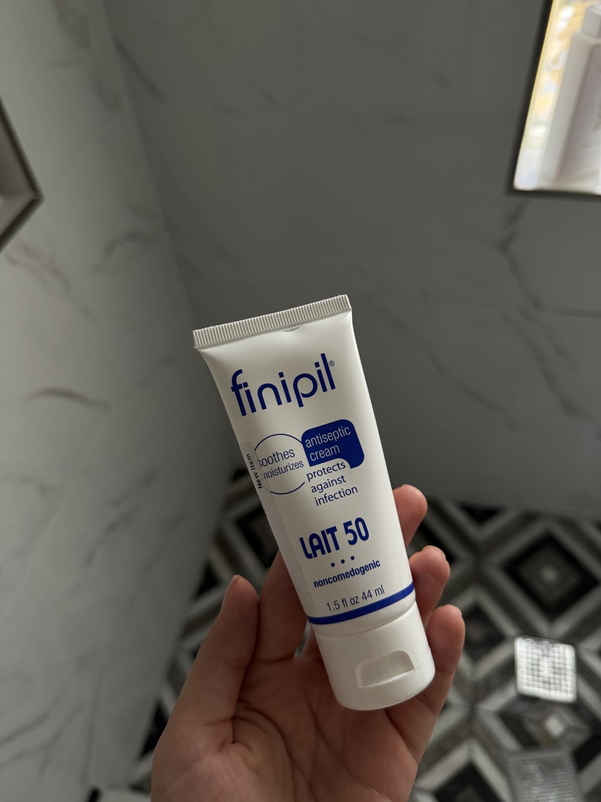 Customer holding a Finipil Lait 50 1.5 oz tube in a bathroom with marble walls and a patterned tile floor